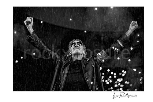 Horizontal black and white image of Roger Waters on stage with both arms raised, wearing dark glasses and a long coat in a dramatic rain-soaked live performance scene with glowing lights behind him