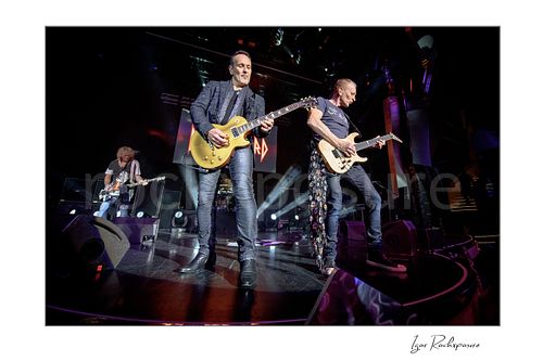 Horizontal color image of Rick Savage, Vivian Campbell, and Phil Collen of Def Leppard performing live on stage under bright concert lighting