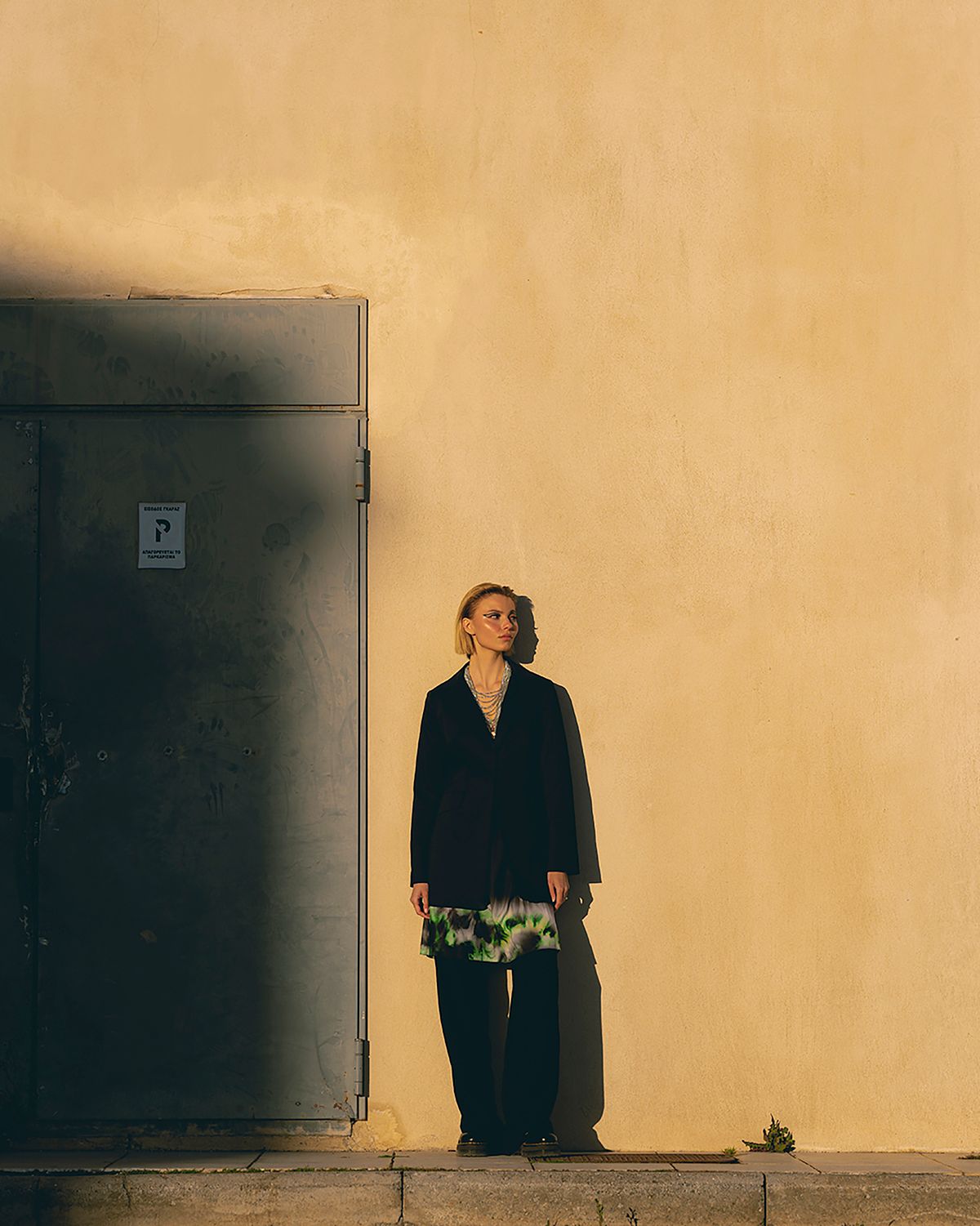 Portrait of a female model wearing a black total outfit with green accents leaning on a yellow wall during sunset light. Photographed by Filianna Karamanli.