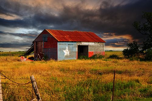 A weathered metal barn painted with the Texas Lone Star flag standing in a golden field under a dark, dramatic storm sky.