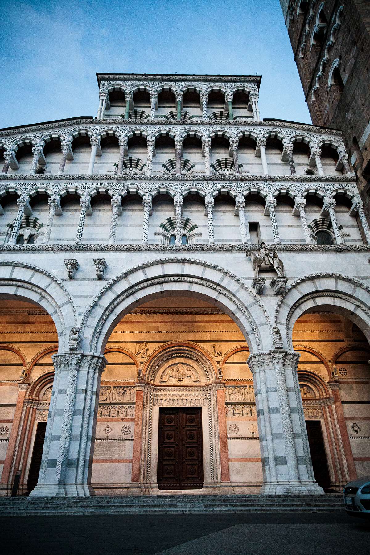Evening view of Lucca Cathedral&rsquo;s ornate stone facade in Italy, showing multiple layers of arches and columns, a large wooden entrance door framed by carved reliefs, and contrasting light and dark stone patterns, softly lit against a clear blue sky with part of the street and a vehicle visible in the foreground.