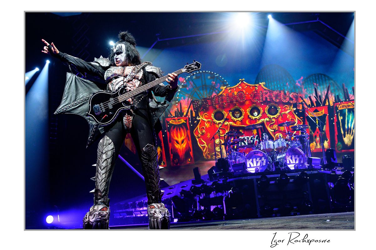 Gene Simmons of KISS performing in a wide power stance with bass guitar under bright white and blue stage lighting with fiery stage visuals behind him
