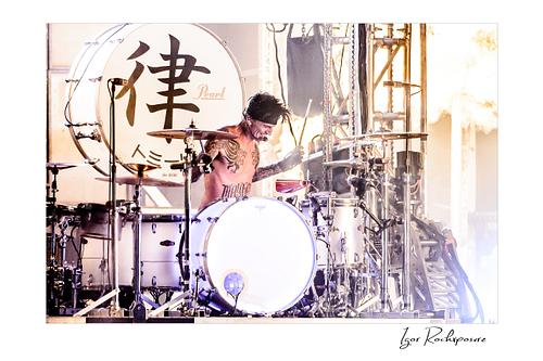 Horizontal color image of Tommy Lee performing at the drum kit during a Mötley Crüe concert, surrounded by cymbals and bright backlight