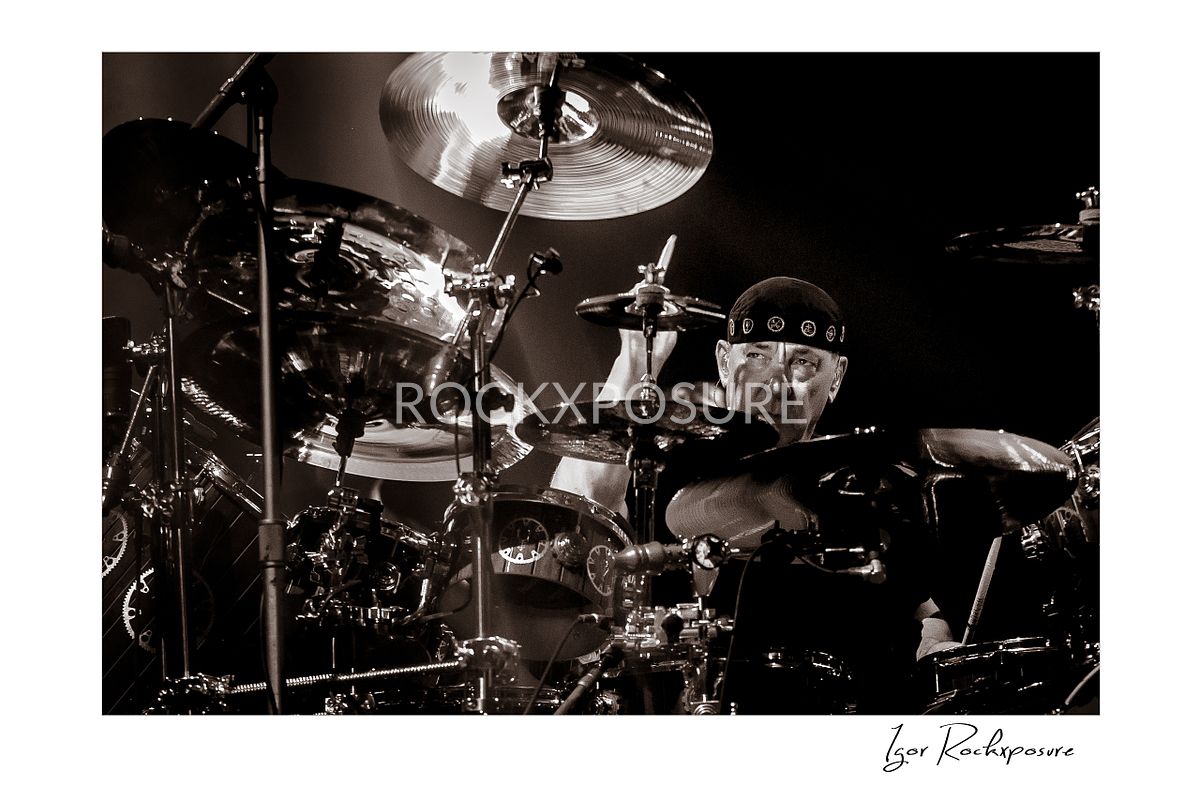 Horizontal concert photography of Neil Peart playing a large drum kit in black and white photography under stage lighting