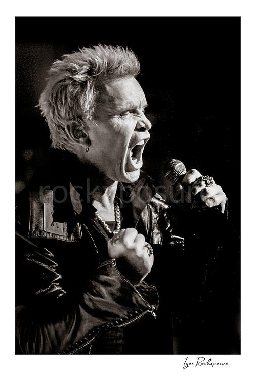 Vertical black and white image of Billy Idol singing into a microphone in a tight high-energy live performance close-up