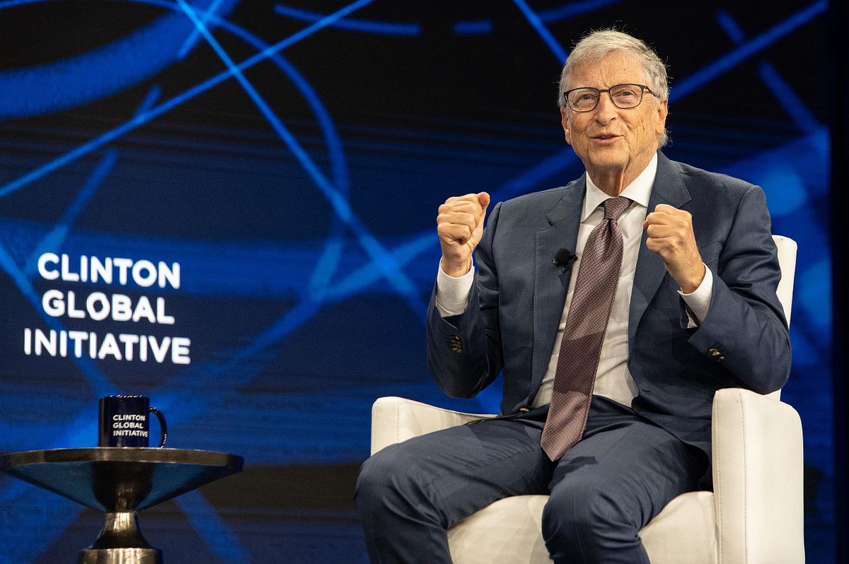 Keynote speaker gestures during a global conference session, with event branding on a blue stage backdrop&mdash;strong presence and authority.