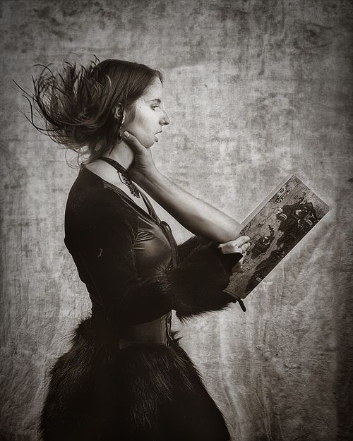 Portrait artistique en noir et blanc d&rsquo;une femme en costume tenant un livre dont une main semble &eacute;merger, la saisissant &agrave; la gorge, &eacute;voquant une absorption surr&eacute;aliste par la fiction &mdash; photographie fine art par Pascal Yennis, Namur, Belgique.