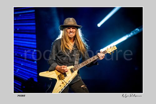Horizontal color image of C.C. DeVille performing with electric guitar during a Poison concert under bright blue stage light