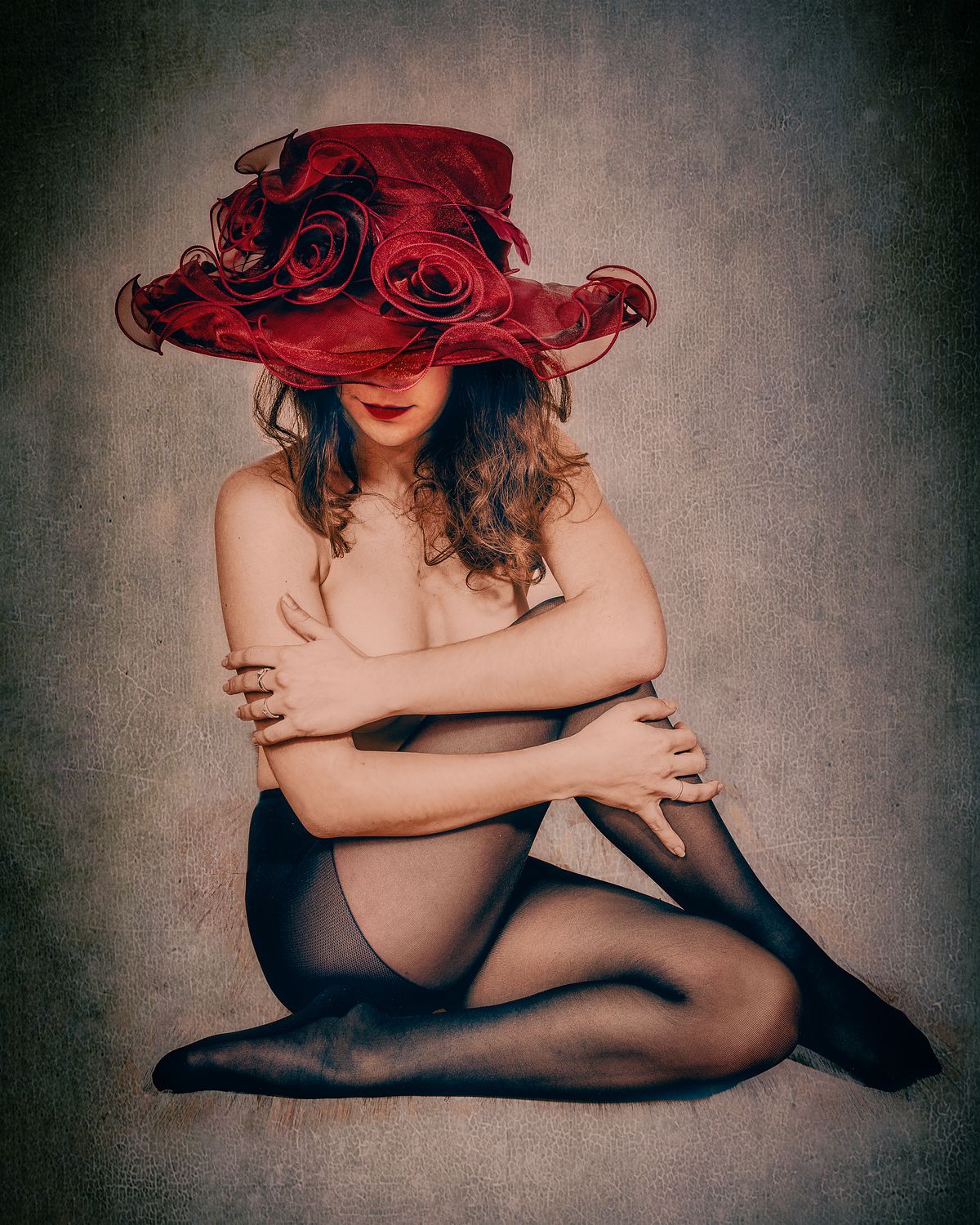 Photographie fine art d’une femme assise nue en collants noirs, coiffée d’un large chapeau rouge à fleurs, dans une posture repliée mêlant pudeur et sensualité — œuvre de Pascal Yennis, Namur, Belgique.