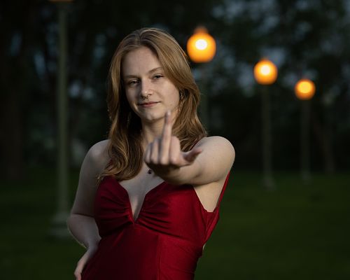 Graduating student in a red dress in front of a row of lights giving the middle finger
