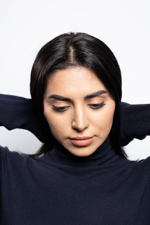 Närbild av kvinna med mörkt hår och händerna bakom huvudet, ögon nedslagna mot vit bakgrund