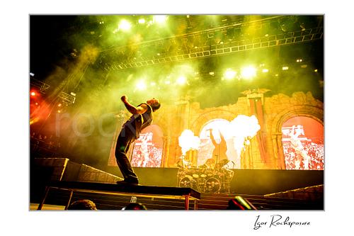 Horizontal color image of M. Shadows of Avenged Sevenfold leaning back on stage with fire bursts, stage smoke, and vivid green concert lights