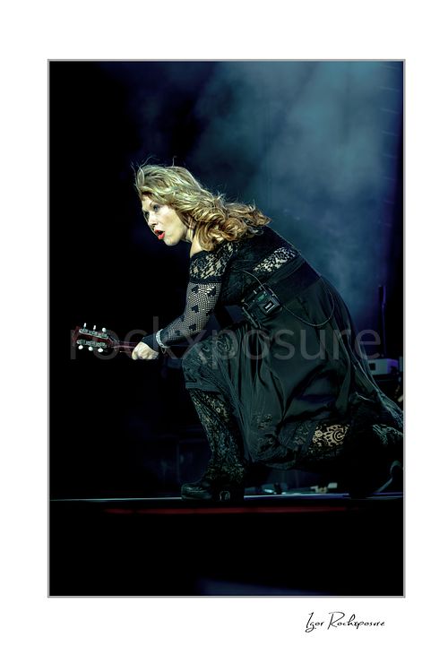 Vertical color image of Nancy Wilson crouching on stage with electric guitar during a Heart concert, framed by blue stage smoke