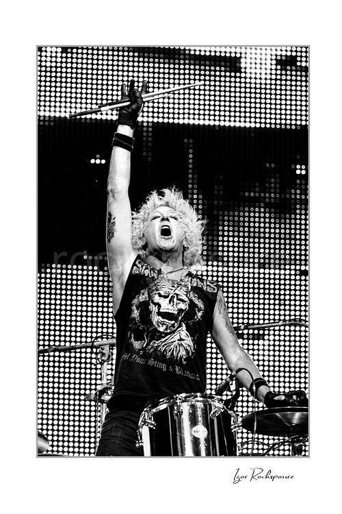 Vertical black and white image of James Kottak of Scorpions behind the drums with raised drumstick and wild hair under a bright LED backdrop