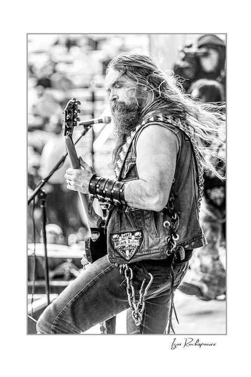 Black and white vertical shot of Zakk Wylde with long hair and a beard, wearing a denim vest, playing guitar and singing into a microphone.