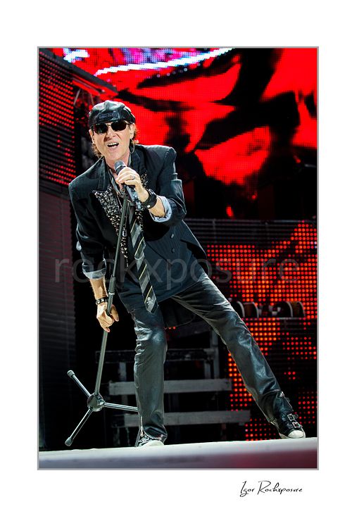 Vertical color image of Klaus Meine of Scorpions singing live with microphone stand, sunglasses, and vivid red stage screens