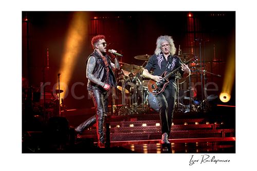 Horizontal color image of Adam Lambert and Brian May of Queen performing onstage together with the drummer visible behind them under warm red concert lighting
