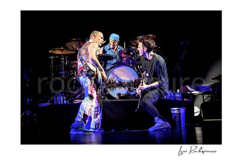 Horizontal color image of Flea, Chad Smith, and Josh Klinghoffer of Red Hot Chili Peppers performing live around the drum kit, with Smith at center while Flea and Klinghoffer face each other on either side