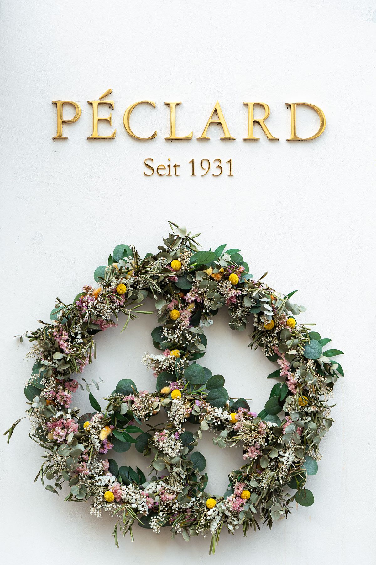 A circular wildflower wreath with eucalyptus leaves, white baby's breath, pink flowers, and yellow billy buttons hanging on a white wall beneath golden 'PECLARD Seit 1931' lettering at a historic Swiss shop in Zurich.