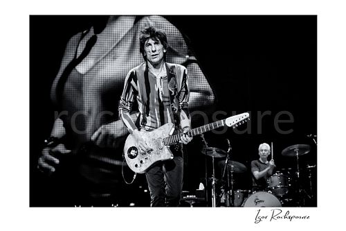 Horizontal black and white image of Ronnie Wood and Charlie Watts of The Rolling Stones performing on stage with electric guitar and drums