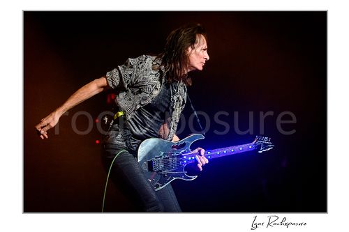 Horizontal color image of Steve Vai playing guitar in side profile with an outstretched arm under warm stage lighting and glowing fret markers