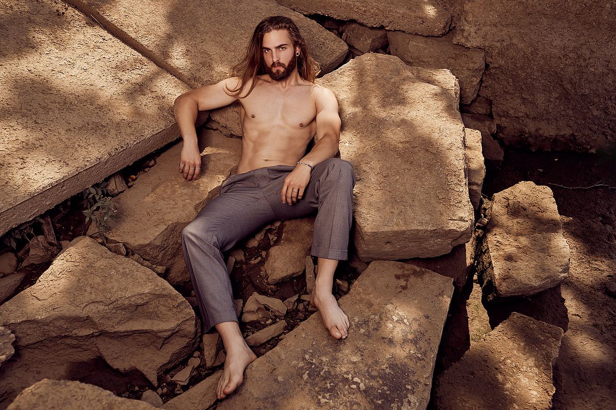 Shirtless male model posed for an editorial portrait outdoors on broken rocks lit with one light and balanced with natural light
