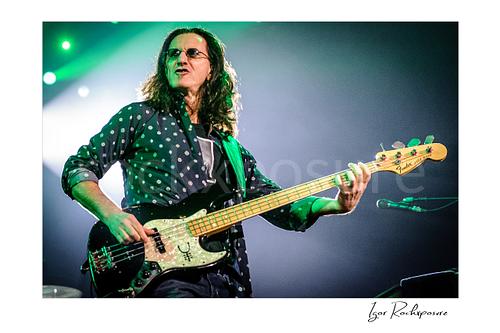 Horizontal color image of Geddy Lee performing with his bass guitar during a Rush concert under green stage light