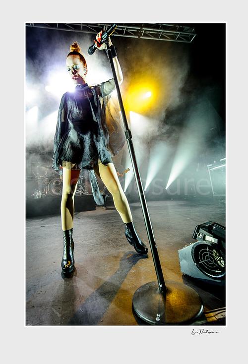 Vertical color image of Shirley Manson standing with a microphone stand during a Garbage concert, framed by stage smoke and bright white and yellow lights