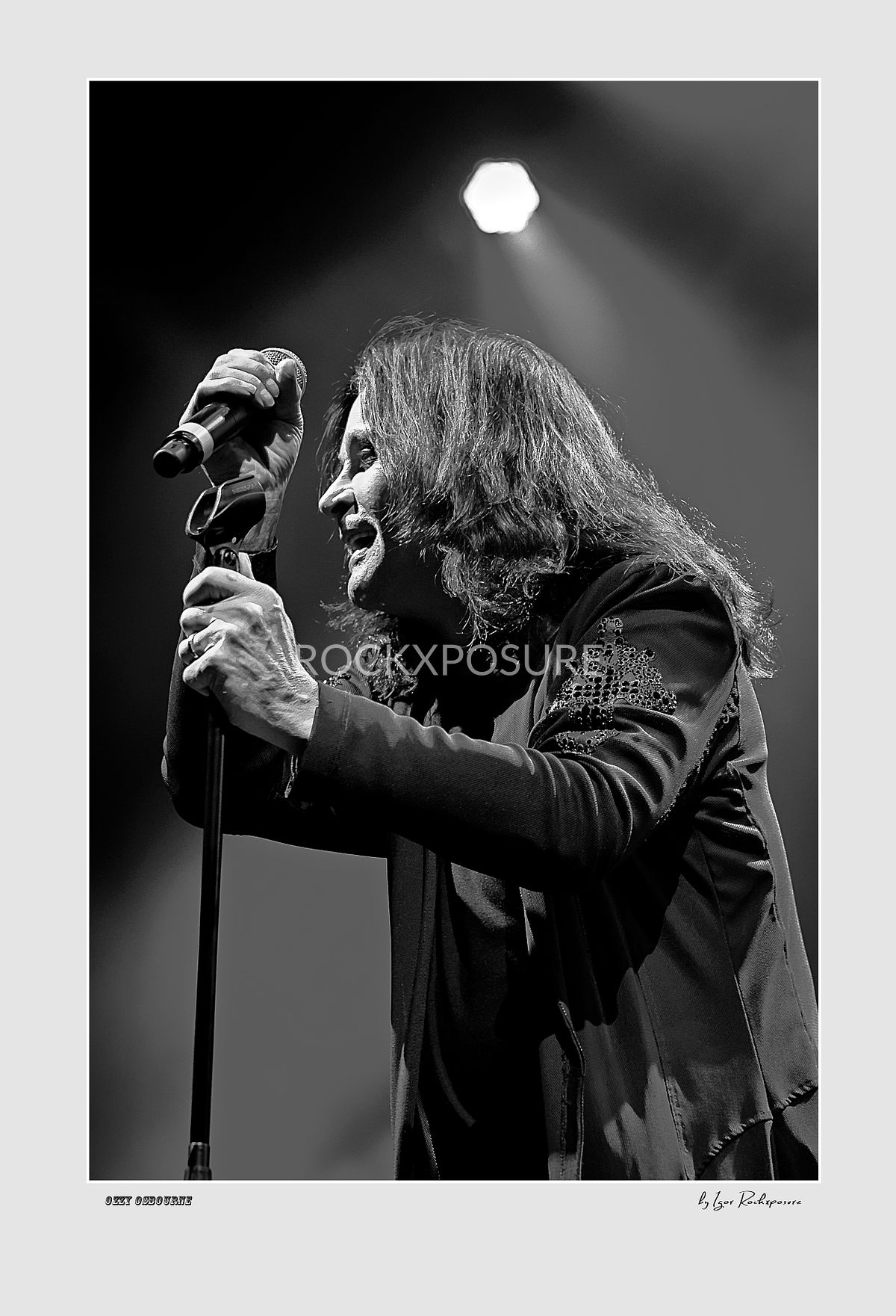 Black and white portrait of Ozzy Osbourne of Black Sabbath leaning toward a microphone stand under dramatic stage lighting