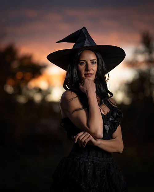 A witch looks at the camera with a colourful sunset behind her.