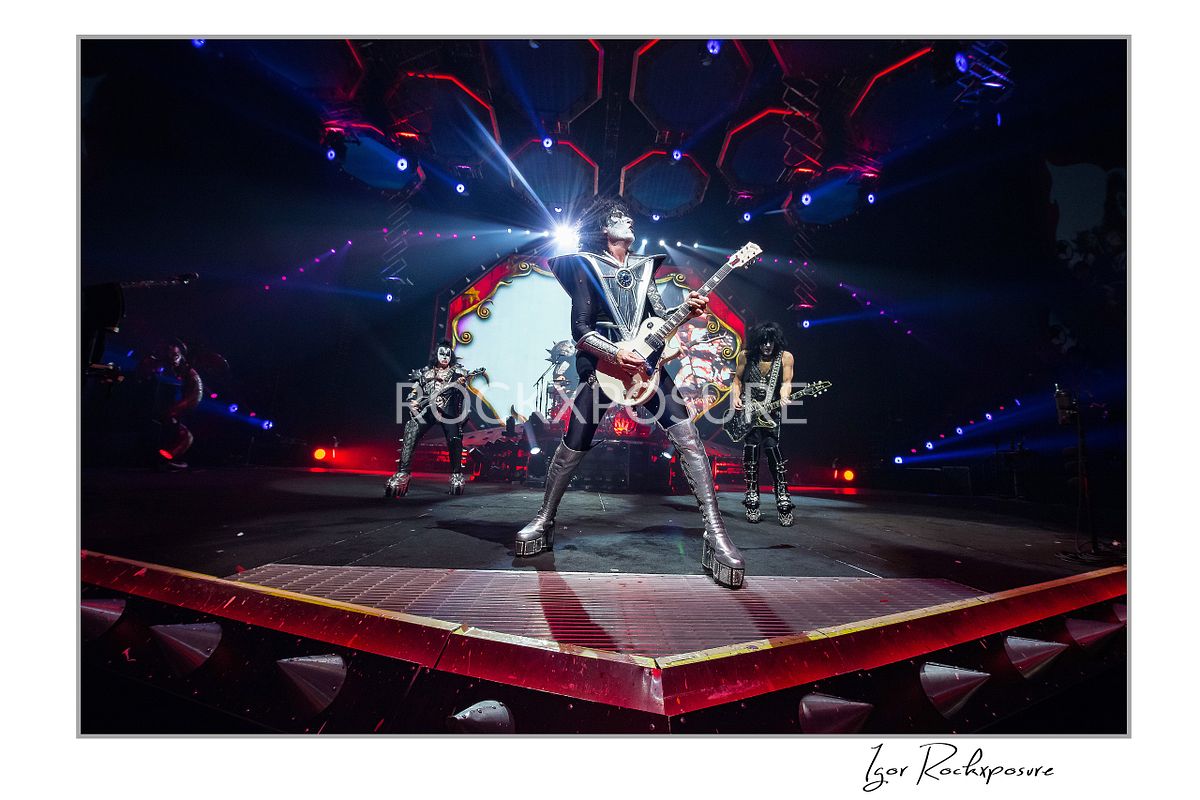 Dramatic horizontal concert photography of Tommy Thayer from KISS in a wide Spaceman stage stance playing electric guitar under intense red and white arena stage lighting with bright stage effects behind him