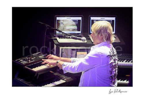 Horizontal color image of Geoff Downes of Yes performing at the keyboard rig with multiple synthesizers and screens during a live performance