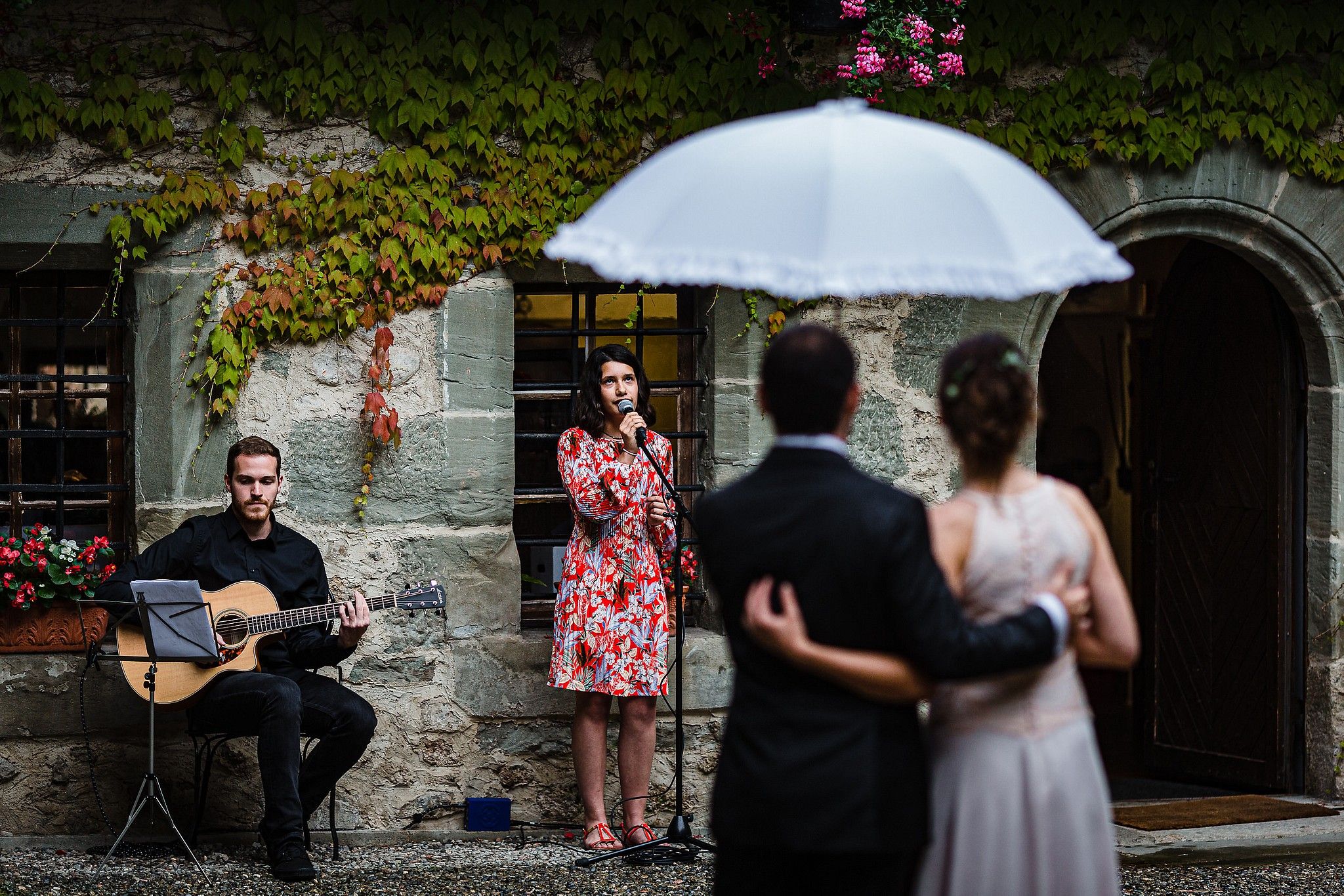 Couple de mari&eacute;s sous un parapluie pendant une averse qui &eacute;coute une enfant chanter captur&eacute; par S&eacute;bastien CLAVEL photographe de Mariage &agrave; Lyon et Gen&egrave;ve