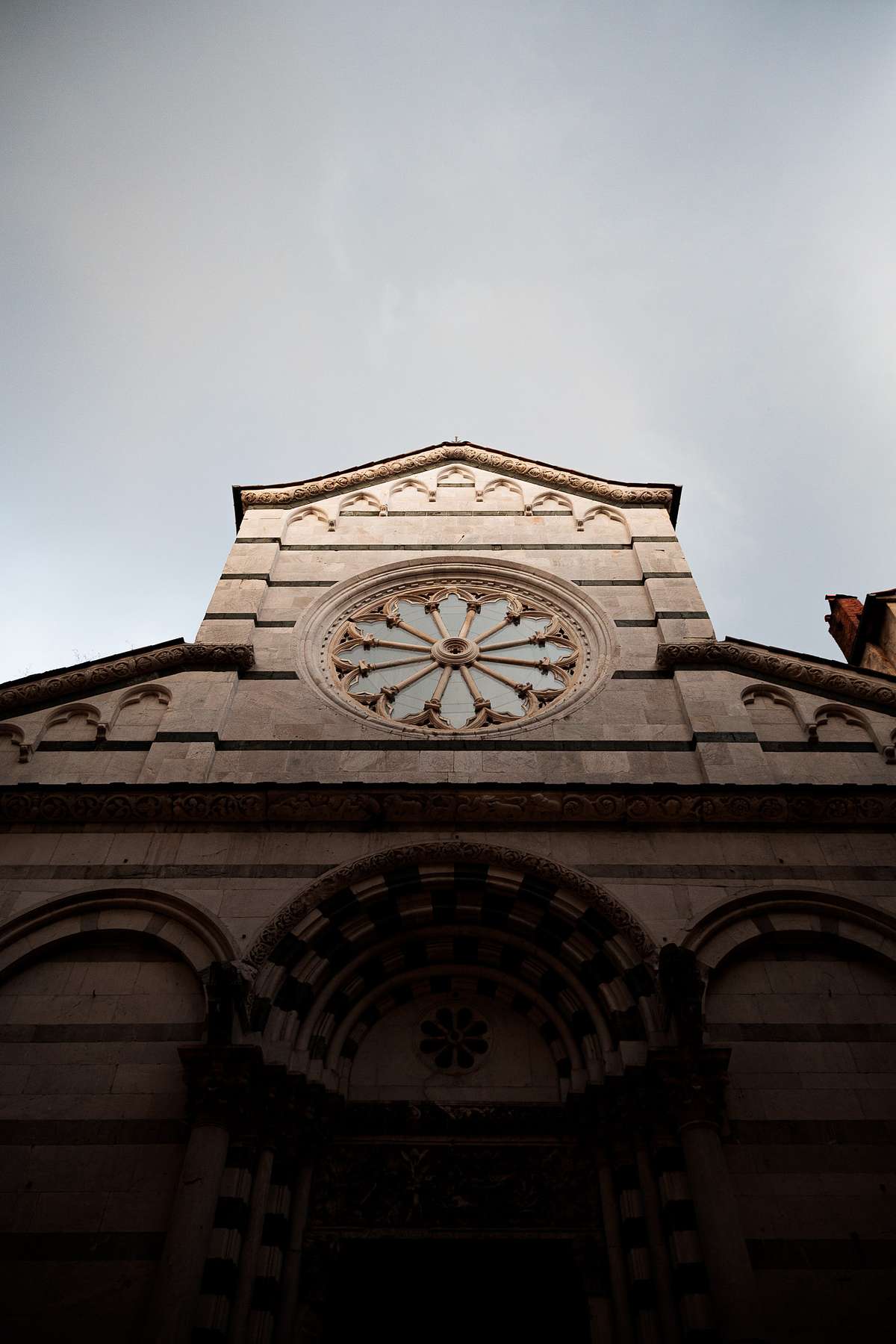 Low-angle photo of a historic Tuscan church facade with light stone stripes, an ornate circular window and a peaked roof, shot under an overcast sky creating a moody atmosphere.