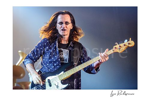 Horizontal color image of Geddy Lee performing with his bass guitar during a Rush concert, with flowing hair and bright stage light