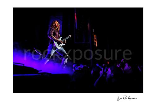 Horizontal color image of Kiko Loureiro of Megadeth playing guitar on stage above purple smoke with fans visible below