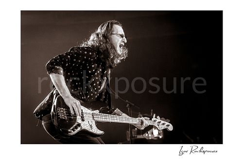 Horizontal black and white image of Geddy Lee performing with his bass guitar during a Rush concert in a dramatic side-profile stage moment