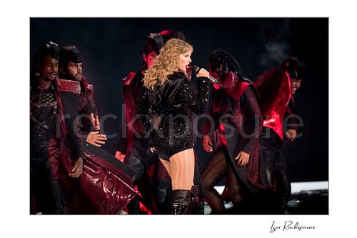 Horizontal color image of Taylor Swift singing on stage in profile with dancers and red concert lighting