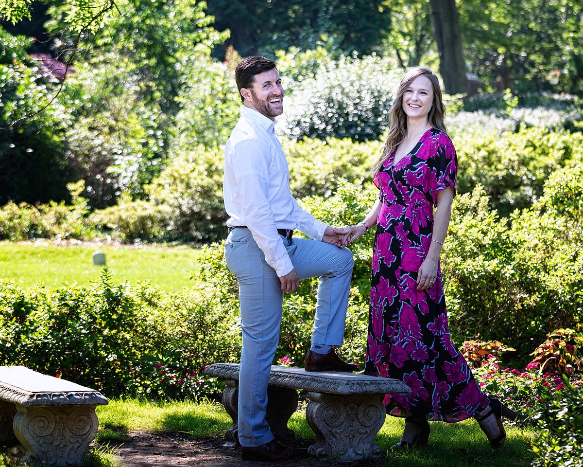 couple in a park on a sunny day, she is wearing a colorful dress and he is wearing jeans and a white shirt. his one leg is propped up on a bench