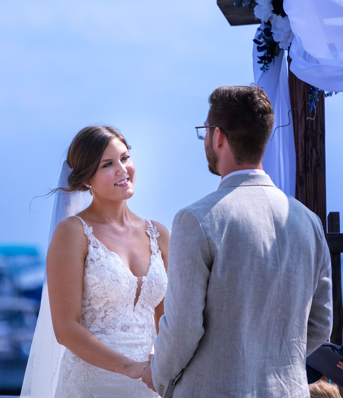 a bride is smiling at looking at the groom during the wedding ceremony on the terrace at hyatt, dewey beach, sussex county, de. she is wearing a white laced dress and a veil, he is wearing a grey suite