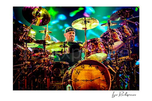 Horizontal color image of Neil Peart performing behind an expansive drum kit during a Rush concert, surrounded by cymbals and vivid multicolored stage light