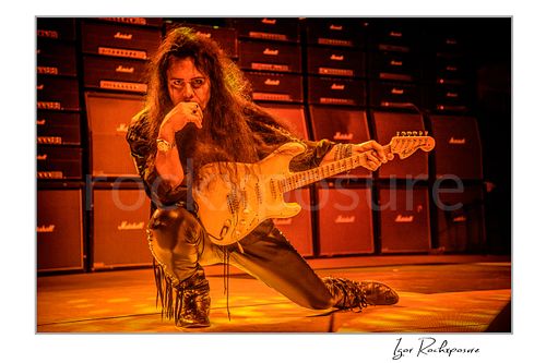 Horizontal color image of Yngwie Malmsteen kneeling on stage with a white guitar under intense warm orange lighting, framed by a wall of Marshall amplifiers behind him