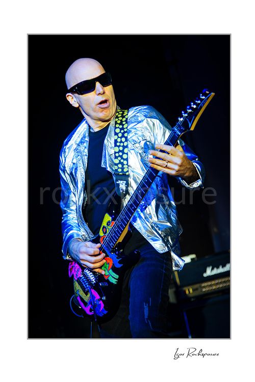 Vertical color image of Joe Satriani performing live in silver jacket and sunglasses with decorated black guitar against a dark stage background