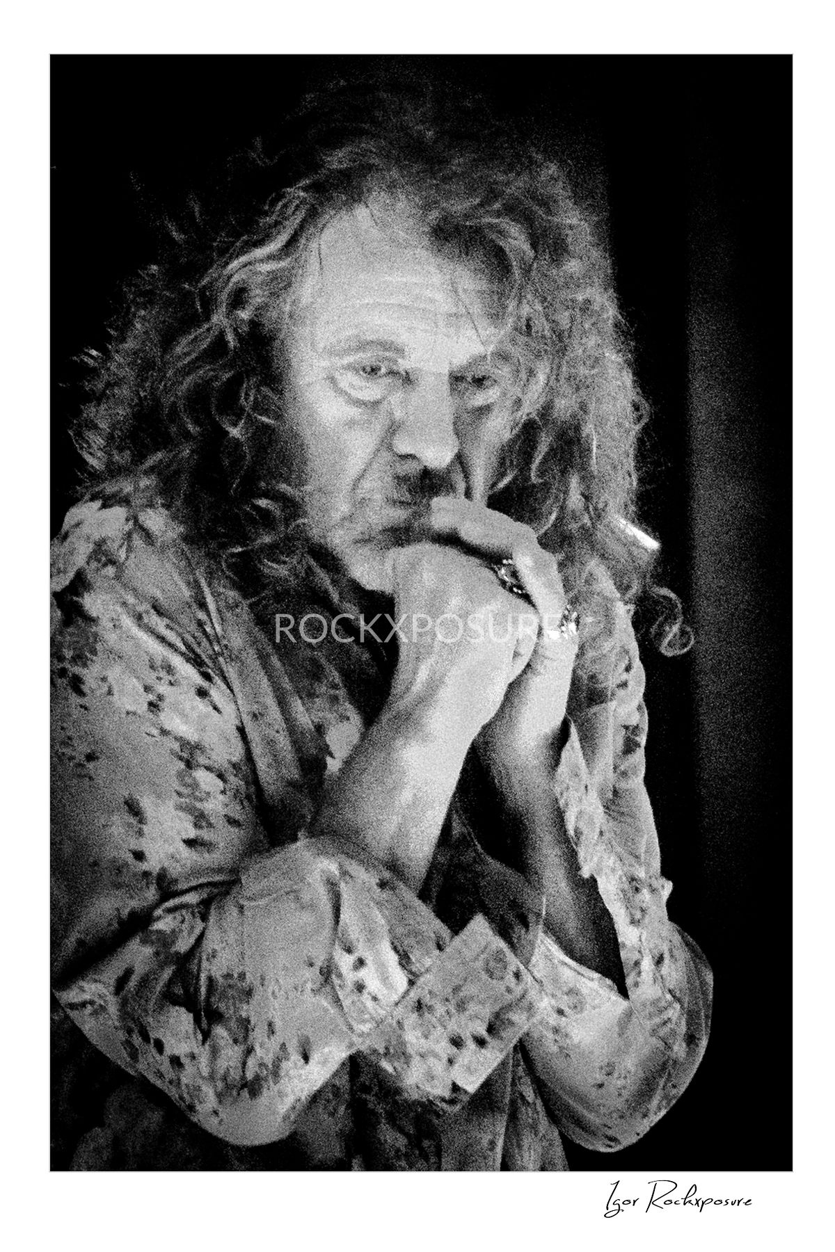 Vertical black and white concert photography portrait of Robert Plant standing with hands clasped together looking down in a deep thoughtful expression under dramatic stage lighting