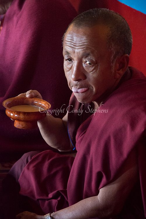 A Nepalese monk dressed in traditional crimson robes sits peacefully, sipping from a wooden bowl, embodying the tranquility of Buddhist monastic life.