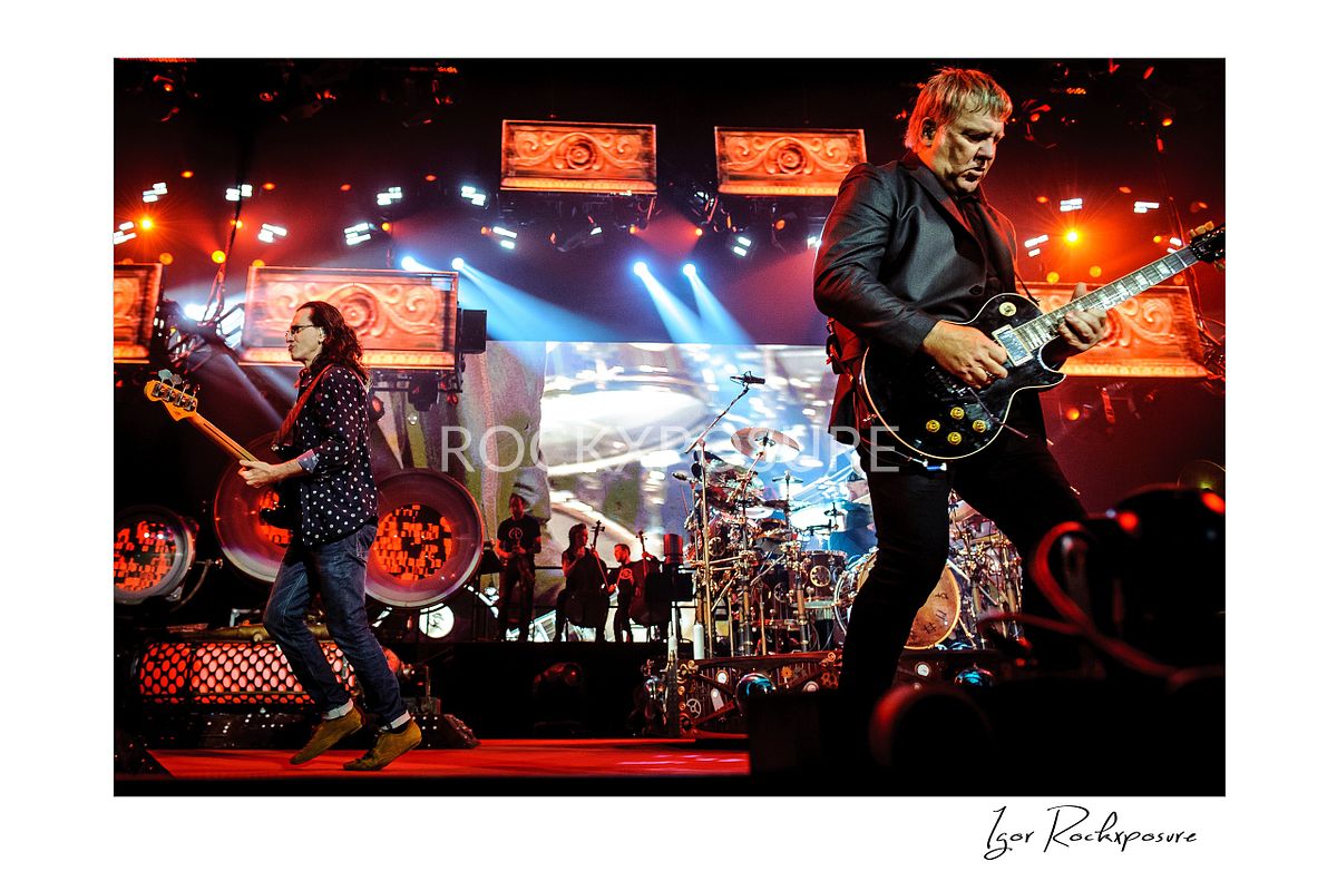 Horizontal concert photography of Rush performing on stage with Alex Lifeson and Geddy Lee under red and blue stage lighting