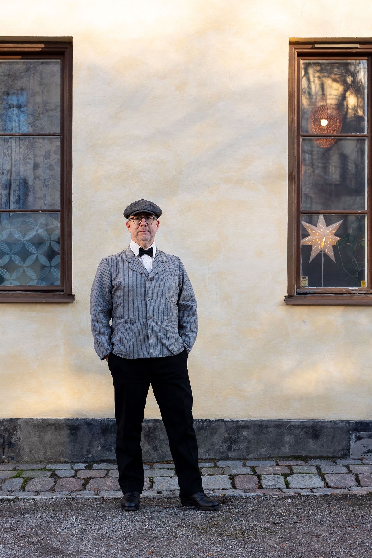 Full-body portrait of a man in a vintage-style outfit standing outside between two windows, photographed in Stockholm by Mats Karlsson.