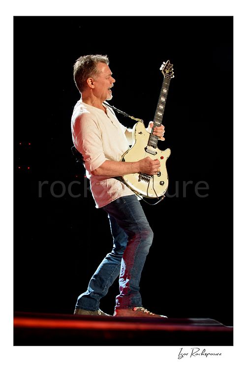 Vertical color image of Eddie Van Halen of Van Halen performing live with a white electric guitar against a dark stage background