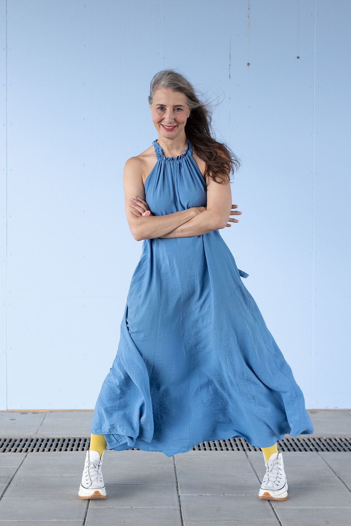 Kvinna i bl&aring; kl&auml;nning och vita sneakers, korslagda armar och vind i h&aring;ret framf&ouml;r ljusbl&aring; v&auml;gg &ndash; redaktionellt milj&ouml;portr&auml;tt i Stockholm.