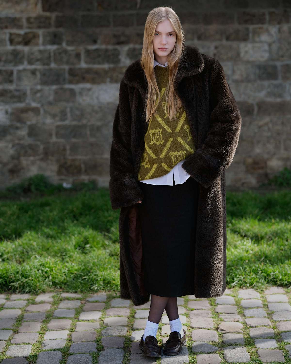 A full frame shot of a woman in a white shirt, lime green sweater, and a fur coat with a brooch, posing for the camera